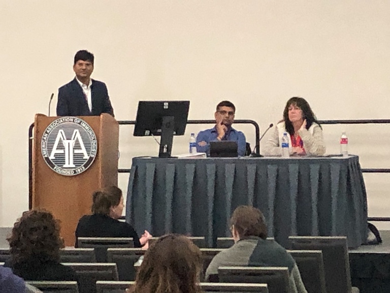 A person at a podium speaking near two people sitting at a table
